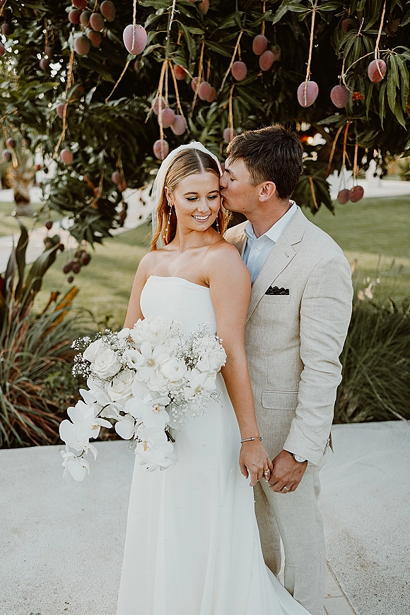 Wedding couple portrait with groom kissing bride’s cheek as she holds a cascading white orchid bouquet beneath a mango tree with hanging fruit