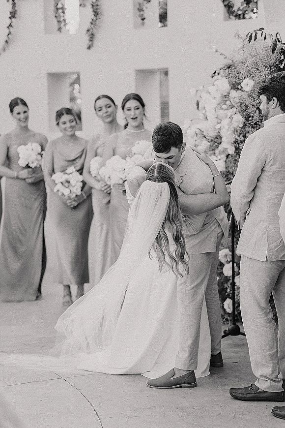 Wedding kiss at the altar as bride and groom embrace beneath a floral arch, bridal veil flowing, wedding party lined up behind outdoors