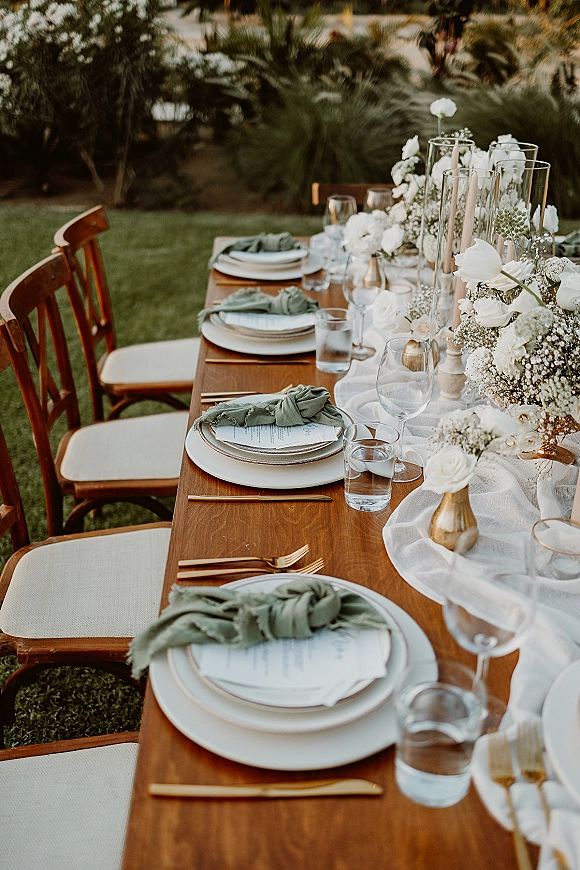 Reception tablescape with an outdoor wedding table set on a wood banquet table, featuring sage napkins, gold flatware, white florals and candles on a garden lawn