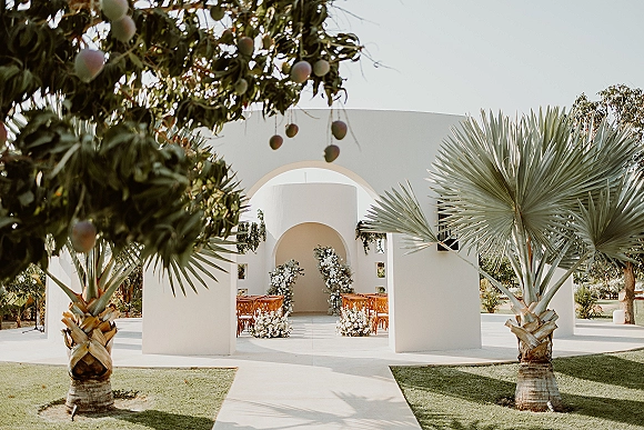Ceremony setup for an outdoor wedding ceremony with wooden chairs and white floral accents, framed by stucco arches and palm trees