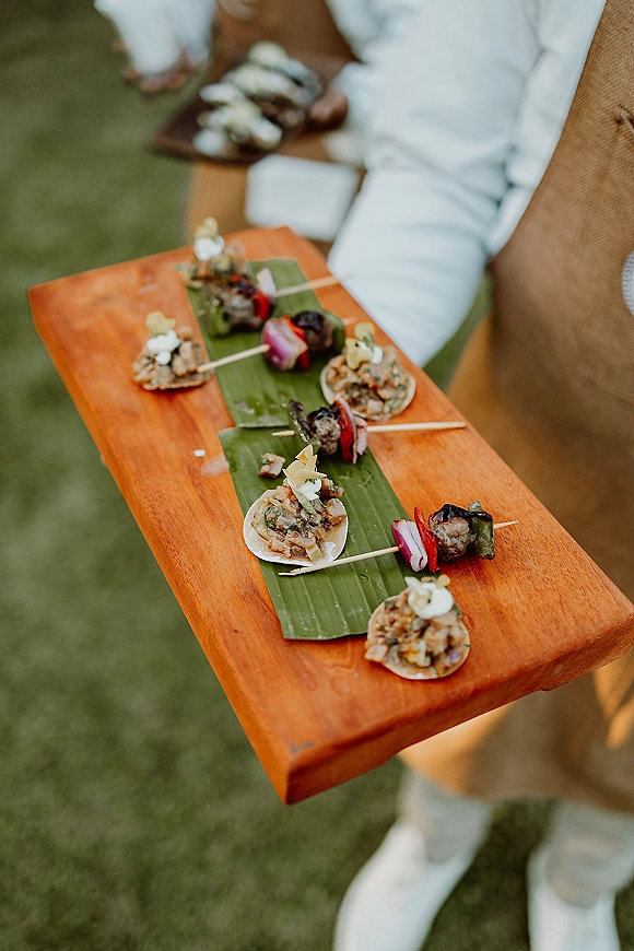 Wedding passed appetizers on a wood serving tray lined with banana leaf, with skewered bite-size hors d'oeuvres held by a server on a lawn