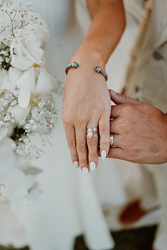 Wedding rings close-up on bride and groom hands, highlighting engagement ring, wedding band stack, white manicure, and baby's breath bouquet.