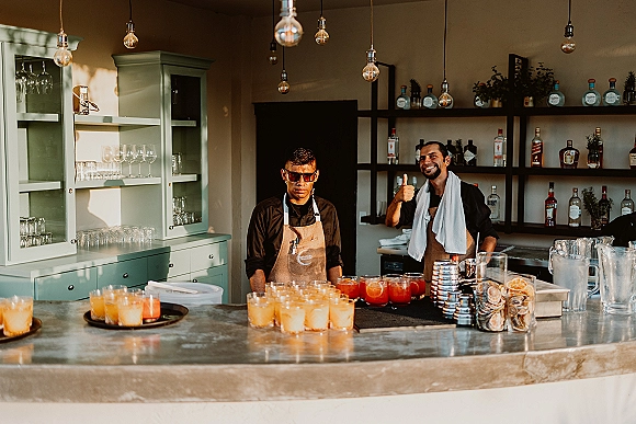 Wedding bar setup with signature cocktail station featuring batch cocktails, bartenders, glassware, liquor bottles, and citrus garnish under pendant bulbs