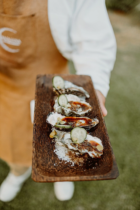 Oyster bar wedding oyster bar platter on a wooden serving board with oysters on the half shell, cucumber slices, sauce, and salt on a lawn
