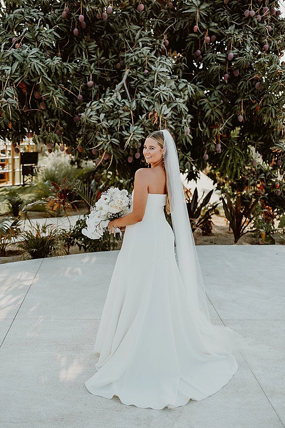Bridal portrait of a bride in a strapless wedding dress with long veil, holding bouquet and looking over shoulder on a tropical patio