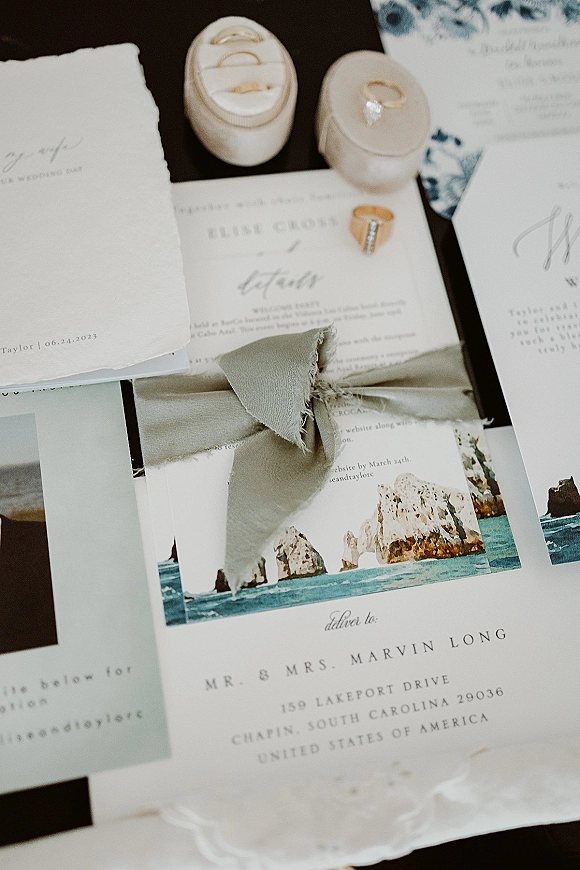 Invitation suite flat lay with silk ribbon wrap, deckle edge cards, photo card, and wedding rings in boxes on a dark tabletop