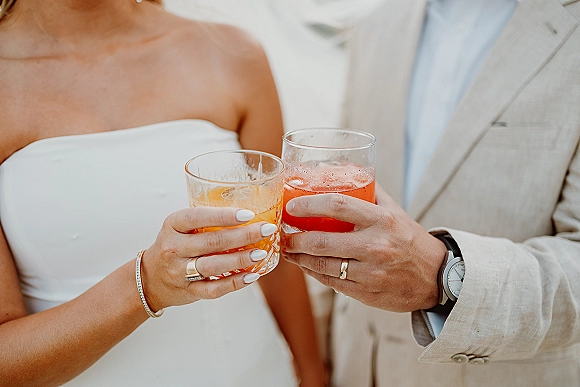 Wedding toast as bride and groom clink cocktail glasses, orange and red drinks shown close up with rings and white manicure on hands