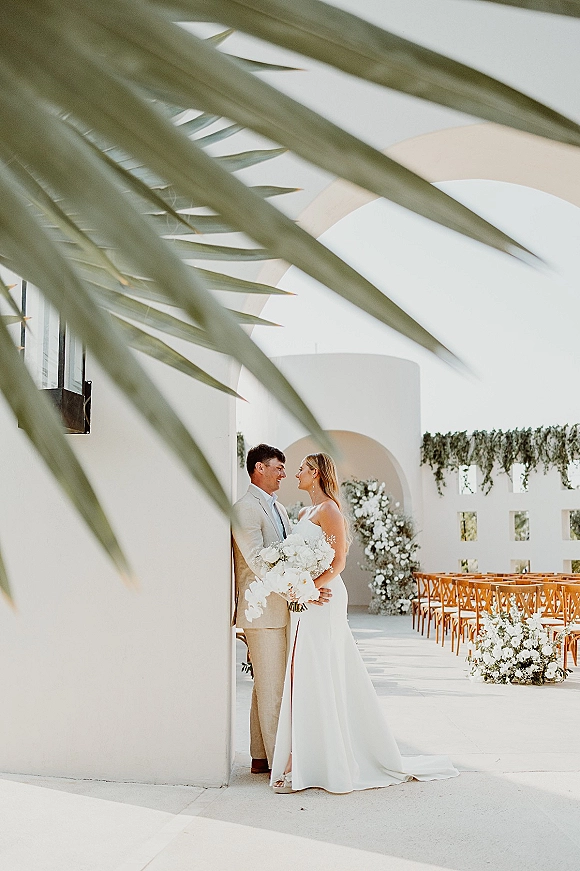 Couple portrait of bride holding white bouquet beside groom in a light suit, framed by a white stucco archway and palm fronds