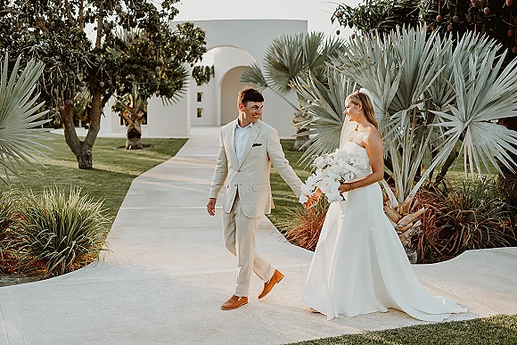 Couple portrait of bride and groom walking hand in hand, bride in veil holding white orchid bouquet on a palm-lined garden walkway