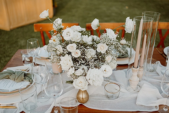 Wedding tablescape with a wedding floral centerpiece of white roses and baby's breath in a brass vase, taper candles and glassware on a lawn
