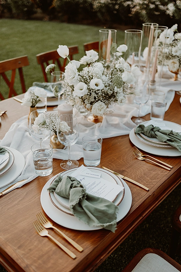 Reception tablescape with an outdoor reception table on a wood farm table, white runner, baby's breath centerpiece, taper and votive candles on lawn