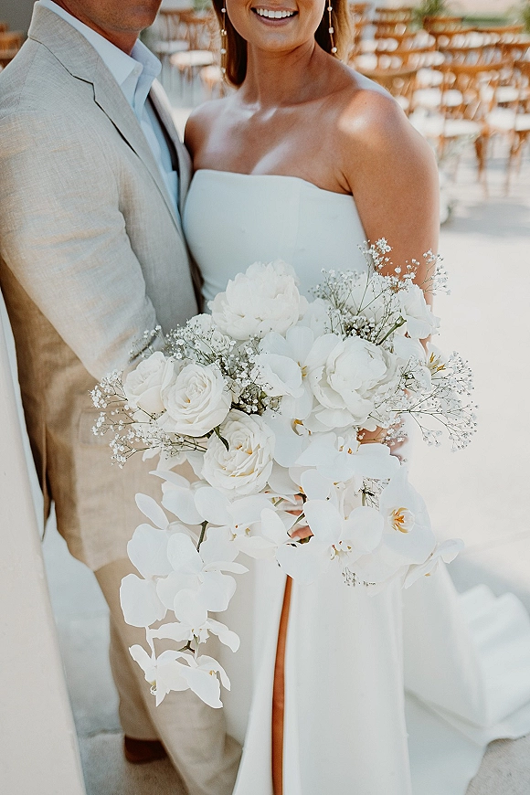 Couple portrait of bride in a strapless wedding dress and groom in a light suit, holding a white orchid bouquet on a sunlit patio