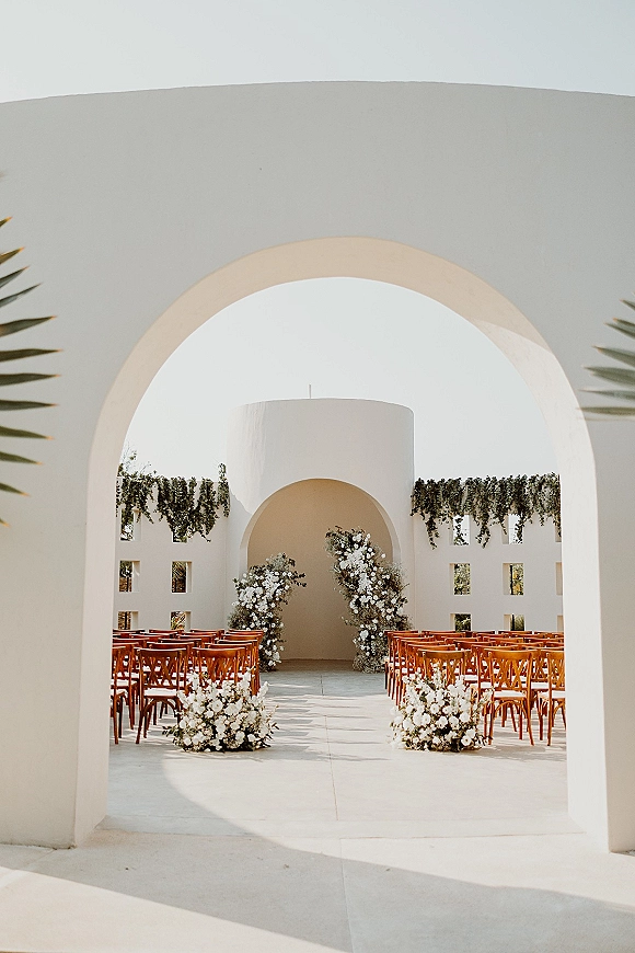 Ceremony setup with wood chairs and floral aisle markers, leading to a greenery-draped arch in a sunlit white stucco courtyard
