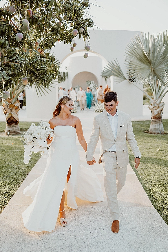 Wedding recessional as bride and groom walking hand in hand, smiling, with cascading white rose bouquet and palm trees by a white arch venue