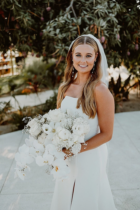 Bridal portrait of a smiling bride holding bouquet of white orchids and baby’s breath, wearing a strapless dress and long veil on a tree-lined walkway