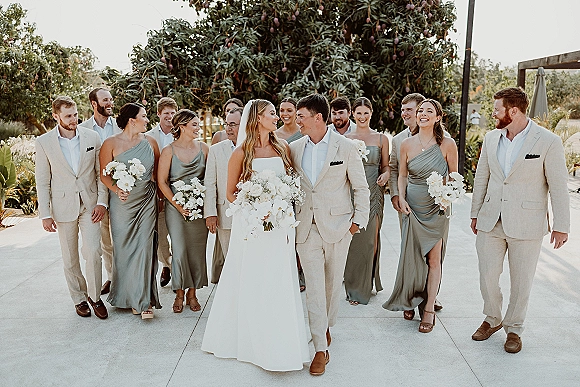 Wedding party portrait with bride in strapless gown and veil, groom in beige suit, and bridesmaids in sage dresses on a leafy patio