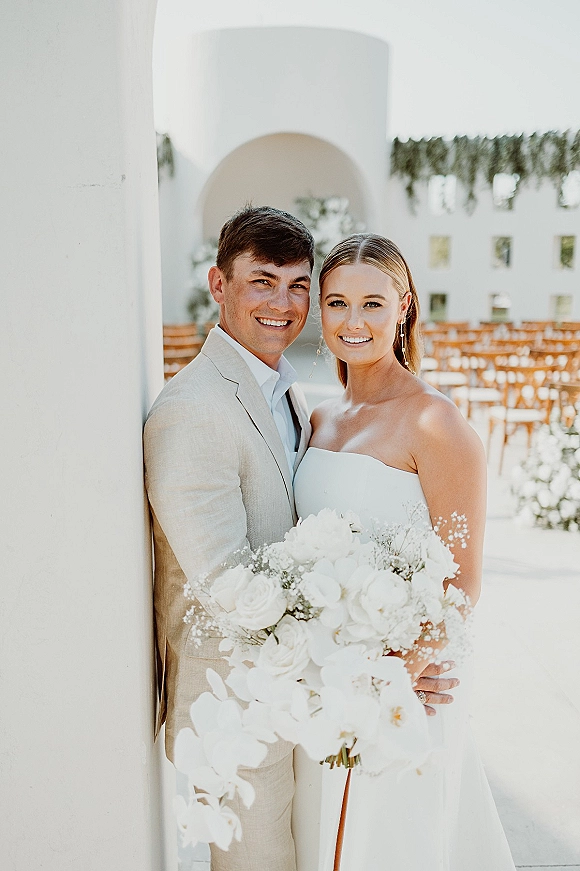 Couple portrait of bride and groom smiling close up, her strapless dress and orchid bouquet against a white stucco arched doorway