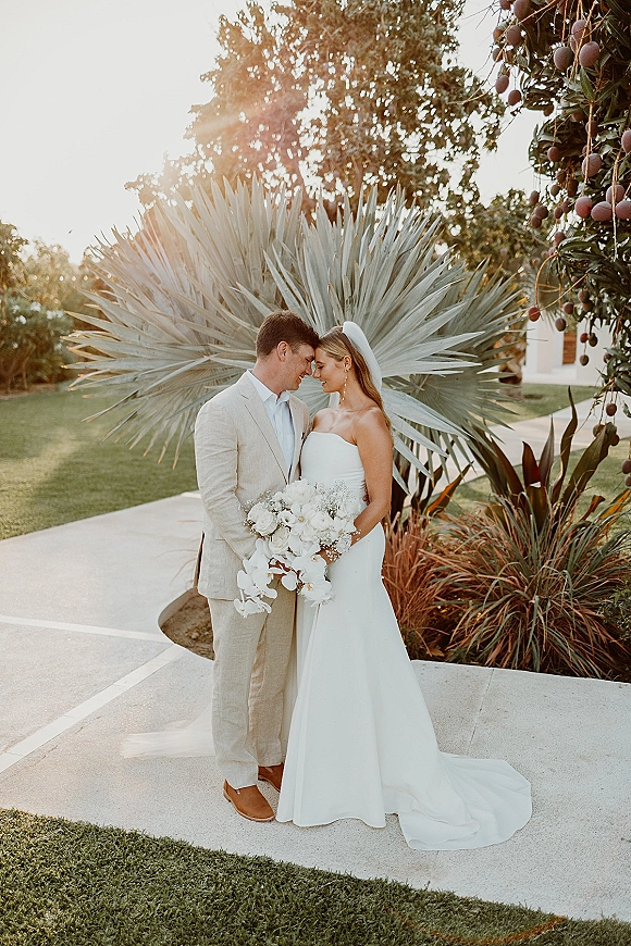 Couple portrait of bride and groom embrace with a forehead touch, bride in strapless gown and veil, holding white orchid bouquet in tropical garden sun flare