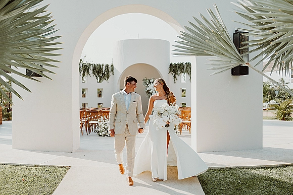 Couple portrait of bride and groom walking hand in hand, her strapless dress and bouquet beside a white stucco archway courtyard with palms