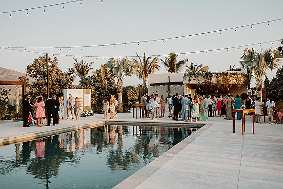 Cocktail hour poolside wedding reception setup with string lights, cocktail tables, and bar station on a patio by palm trees and mountains