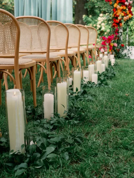Ceremony aisle decor with outdoor ceremony chairs, greenery garland and pillar candles in glass hurricanes on a garden lawn with trees