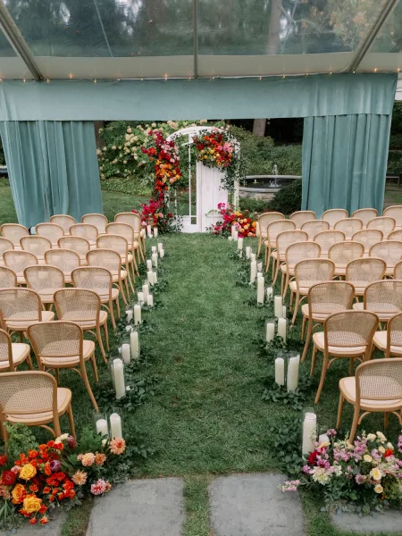 Ceremony setup with a white floral arch and candle-lined outdoor ceremony aisle, framed by teal drapery under a clear-top tent with lights