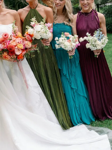 Bridesmaid bouquet photo of colorful hand-tied blooms with ribbon, held by bridesmaids in dresses on a garden lawn by iron fence