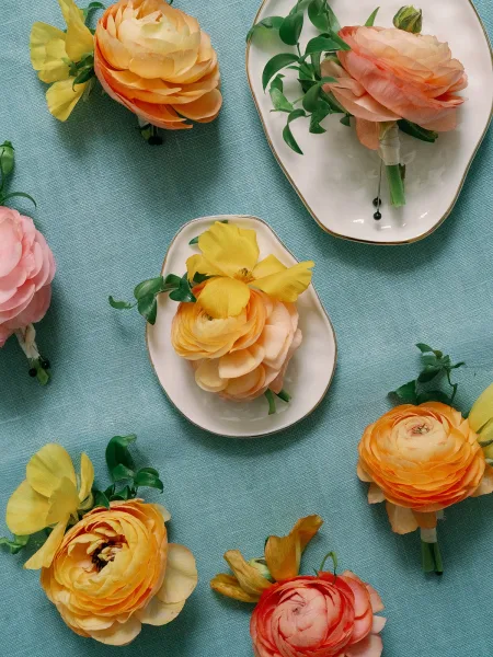 Boutonniere flat lay with ranunculus boutonniere, yellow blooms and greenery, ribbon-wrapped with black pins on blue linen by gold-rim dishes