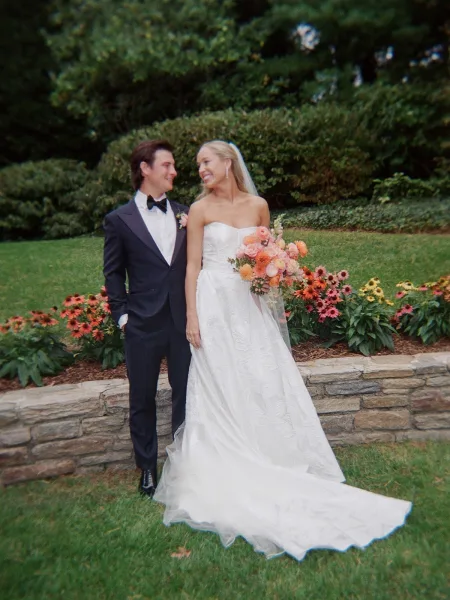Couple portrait of bride holding a colorful bouquet beside groom in tuxedo, smiling on a garden lawn by a stone retaining wall