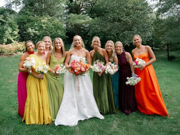 Bride and bridesmaids pose with bouquets in colorful dresses on a garden lawn by trees and a black metal fence, veil flowing