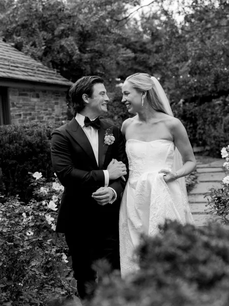 Couple portrait in a black and white wedding portrait style, bride and groom laughing as they walk a stone garden path, her veil flowing