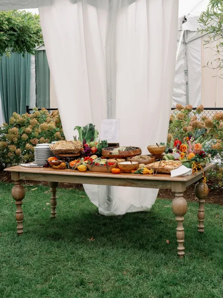 Wedding grazing table with charcuterie table wedding spread of fruit, veggies, breads and mini pumpkins on a wood buffet under white drapery outdoors