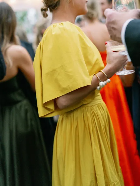 Wedding guest style in a yellow puff sleeve dress holding a champagne coupe, gold bangles and pearl bracelet amid outdoor string lights