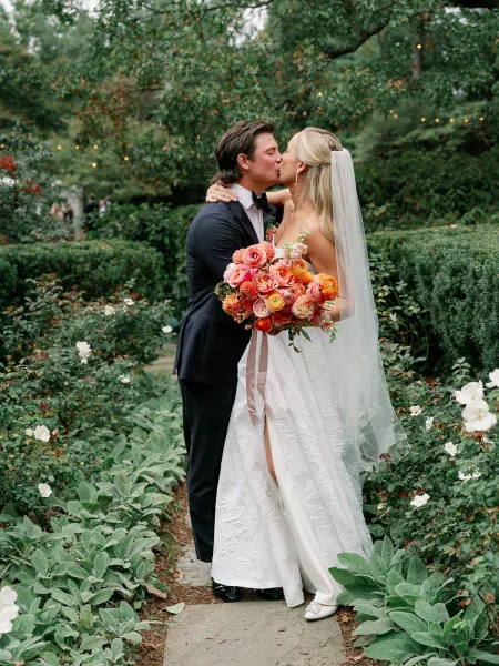 Wedding kiss portrait of bride and groom kissing on a stone garden path, bride holding a coral bouquet, veil flowing past hedges and lights