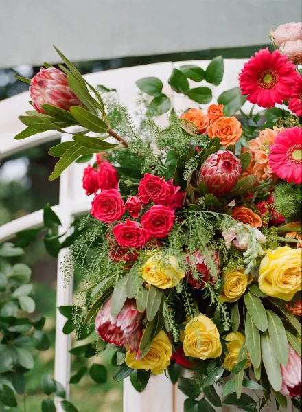 Wedding ceremony flowers with roses, gerbera daisies, and protea blooms draped on a white chair, set against outdoor greenery