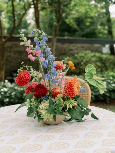Wedding centerpiece of colorful dahlias and delphinium in a low ceramic bowl with greenery, set on a patterned tablecloth outdoors