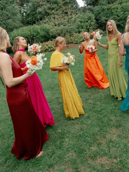 Bridesmaid portraits of bridesmaids holding bouquets in colorful dresses, laughing together on a garden lawn with trees behind