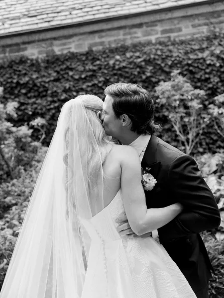 Wedding kiss portrait of bride and groom kissing in a garden, her long veil flowing over a strapless dress as he wears a tuxedo boutonniere