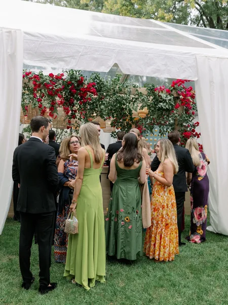 Wedding cocktail hour under a clear top tent with hanging wicker pendant lights, guests in formalwear holding cocktails on a grassy lawn