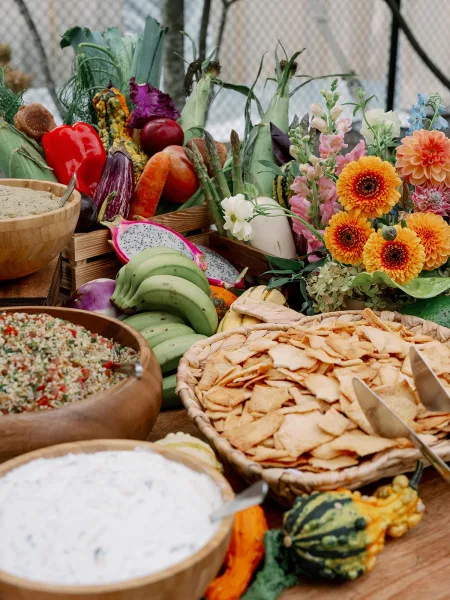 Wedding cocktail hour spread with dips, pita chips, tabbouleh, and colorful produce in wood bowls on a rustic table with floral accent