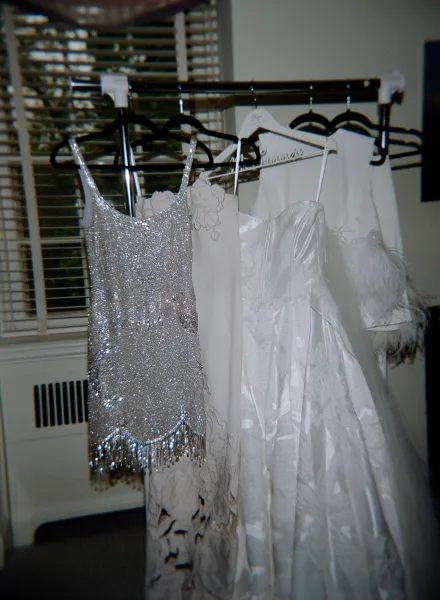 Wedding dresses hanging on a garment rack, with a bridal gown on hanger beside a beaded mini dress in a room with window blinds