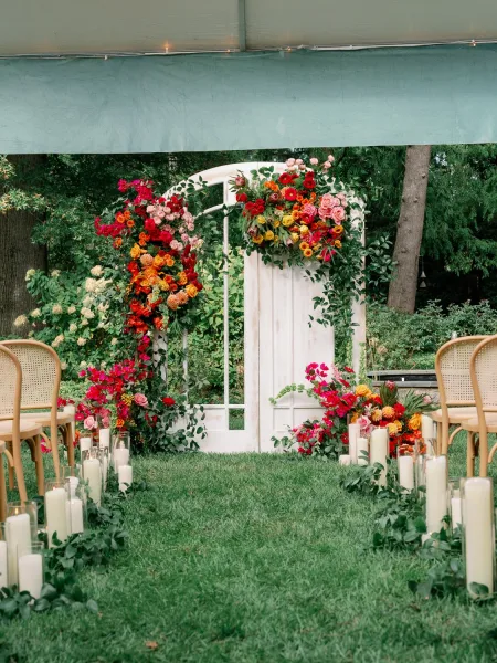 Ceremony backdrop with floral ceremony arch and greenery garland around a white door, candle-lined grass aisle under a tent canopy