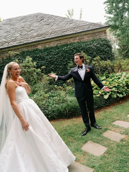 First look moment as bride in a strapless dress and cathedral veil laughs toward groom in black tuxedo by an ivy stone wall