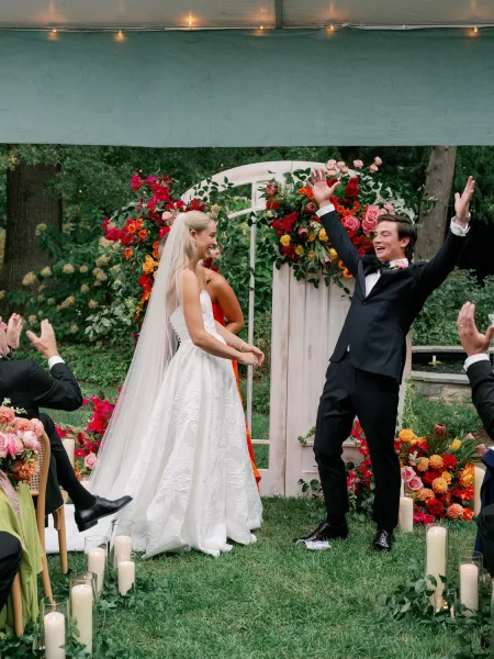 Ceremony moment at an outdoor garden ceremony with bride and groom under a colorful floral arch, veil flowing, aisle candles by tent lights