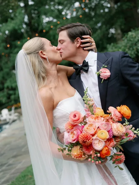 Wedding kiss portrait of bride and groom kissing, her hand on his face showing engagement ring under string lights on an outdoor patio