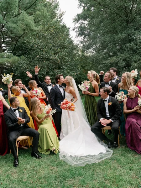 Wedding party portrait of bride and groom kiss as bridesmaids and groomsmen cheer with bouquets on wooden chairs in a garden lawn
