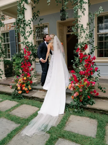 Wedding kiss portrait of bride and groom kissing on stone steps, her long veil trailing behind as they hold hands at a columned porch