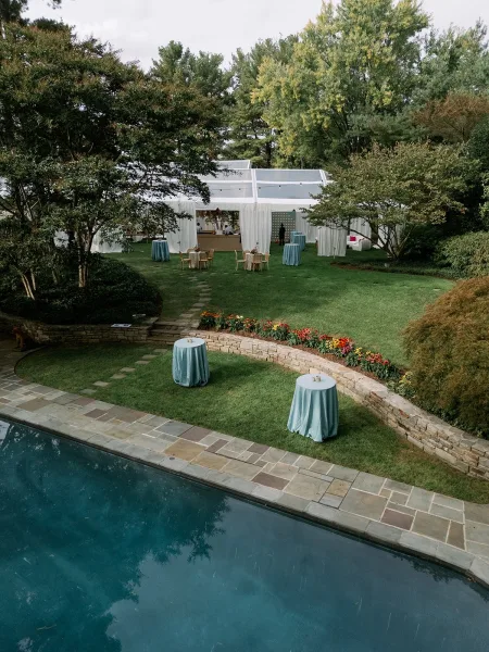 Outdoor reception setup with a white draped tent and string lights, blue linen cocktail tables and wicker lounge seating by the pool
