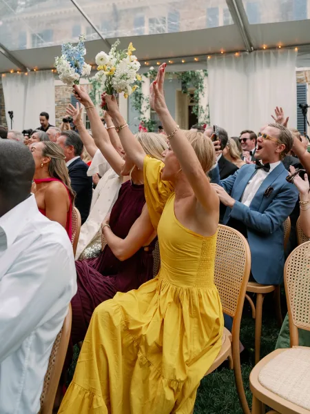 Wedding guest reaction as guests cheer and raise hands, bridesmaids with bouquets in colorful dresses under a clear-top tent with string lights