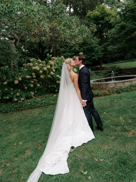 Wedding kiss portrait of bride and groom kissing, her veil trailing behind, on a garden lawn by hydrangeas and a stone fountain